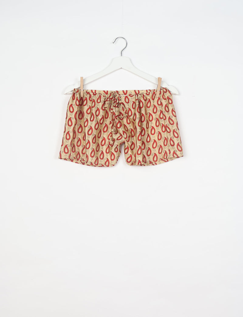 Beige pajama shorts with red paisley pattern and drawstring waist hanging on white hanger against white background