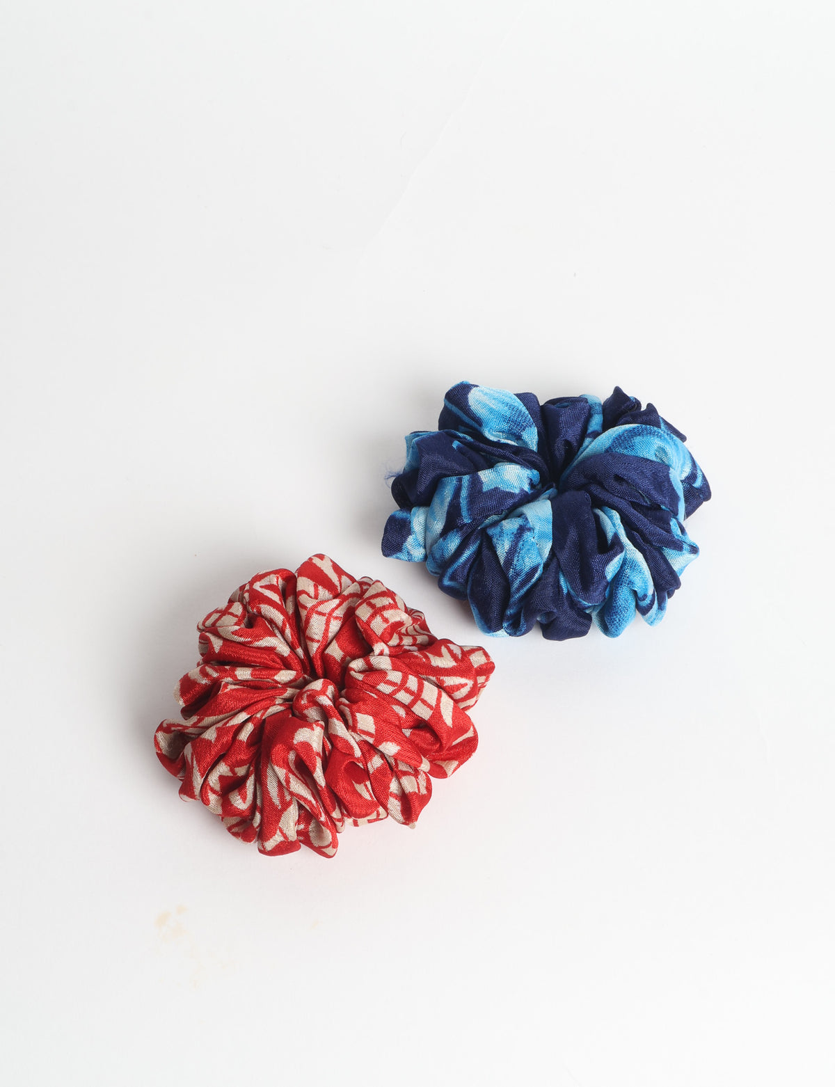 Set of two fabric scrunchies, one turquoise with beige abstract spots, one red with white linear pattern, flat lay on white