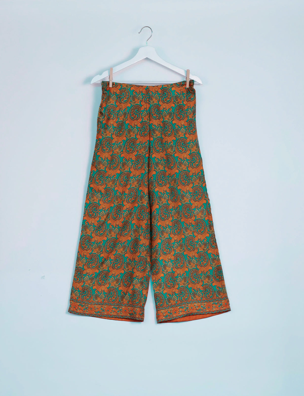 Green and orange paisley patterned wide-leg pants hanging on a white hanger against a light blue background