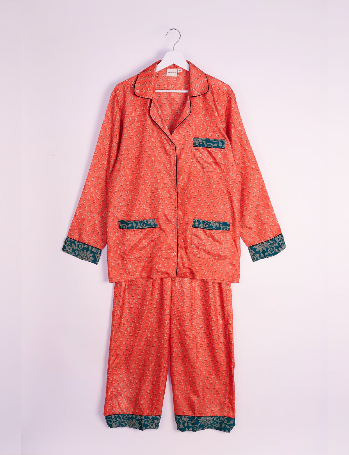 Orange long-sleeve pajama set with micro floral design and contrasting dark green patterned cuffs and pockets, hanging on a white hanger