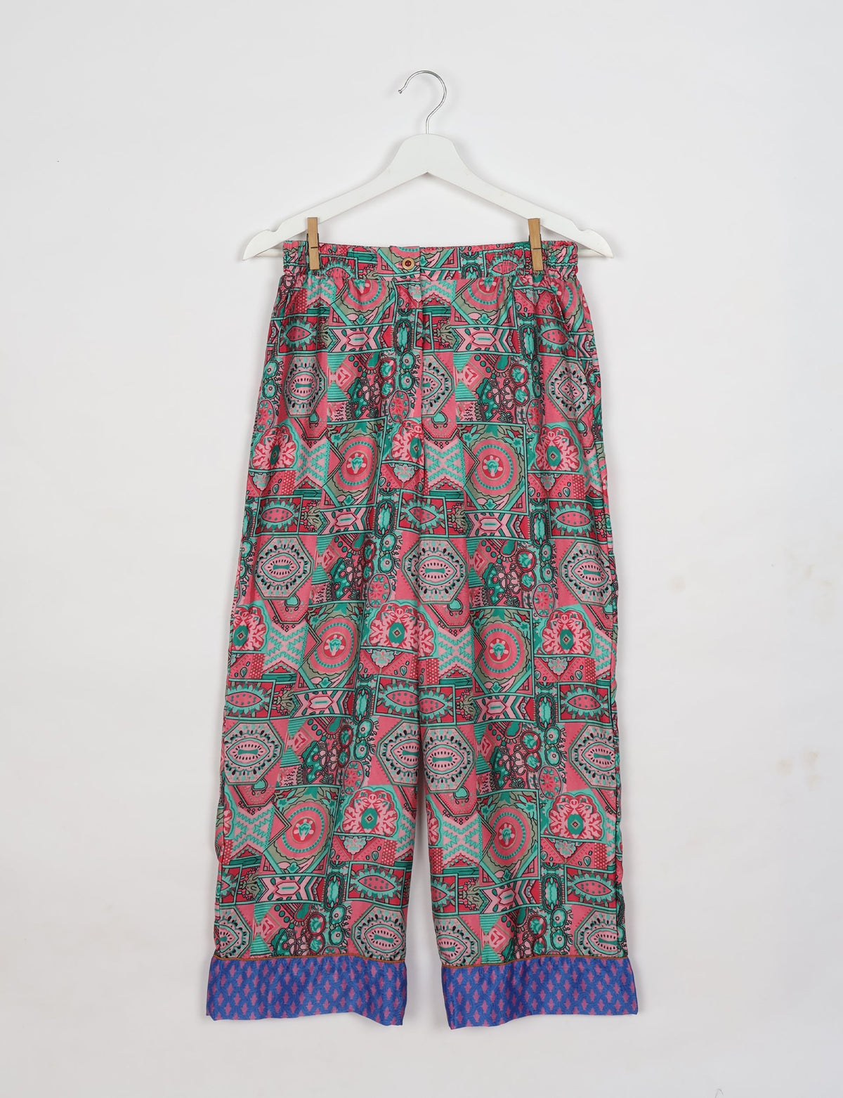 Pink ethnic patterned pajama pants with blue patterned cuffs, hanging on a white hanger against a white background