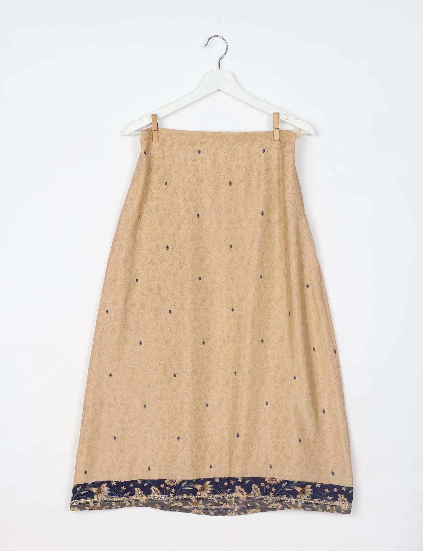 Beige A-line skirt with subtle floral pattern and navy floral border hem, hanging on white hanger with wooden clips