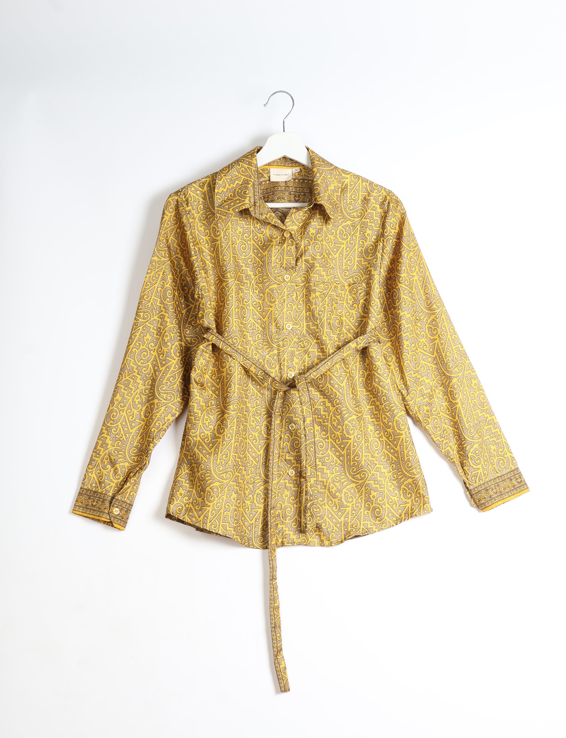 Yellow long-sleeve button-up shirt with intricate paisley pattern and waist tie, hanging on white hanger against white background