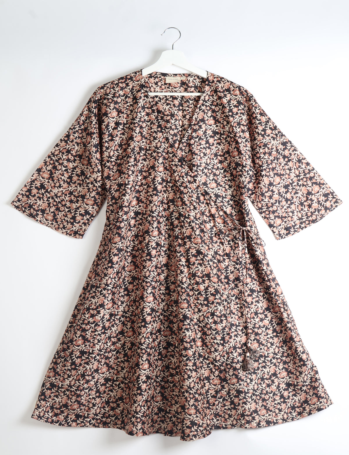 Black coat with pink and beige floral tropical print, wide sleeves, wrap style, and tassel tie detail, hanging on white