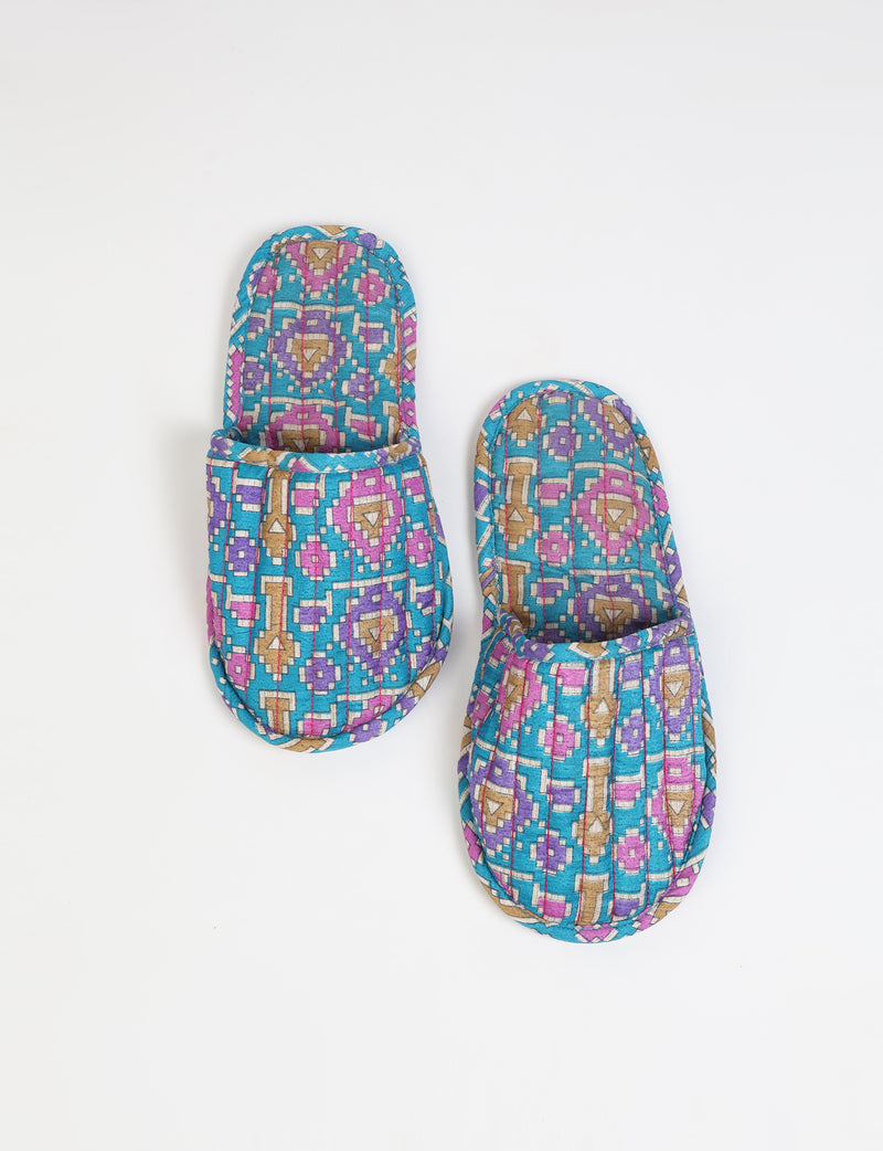 Blue quilted slippers with geometric pattern in purple, tan, and white, shown from top view on white background