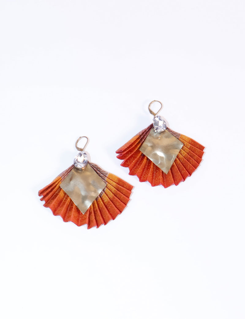 Pair of pleated earrings with orange abstract fabric, gold-tone diamond-shaped metal, and clear teardrop crystals, flat lay view