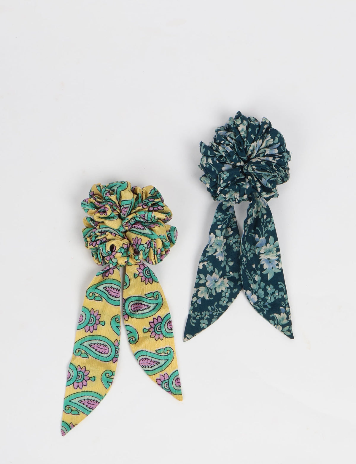 Set of 2 tail scrunchies with attached fabric tails, one yellow with paisley print, one dark blue with floral print, flat lay