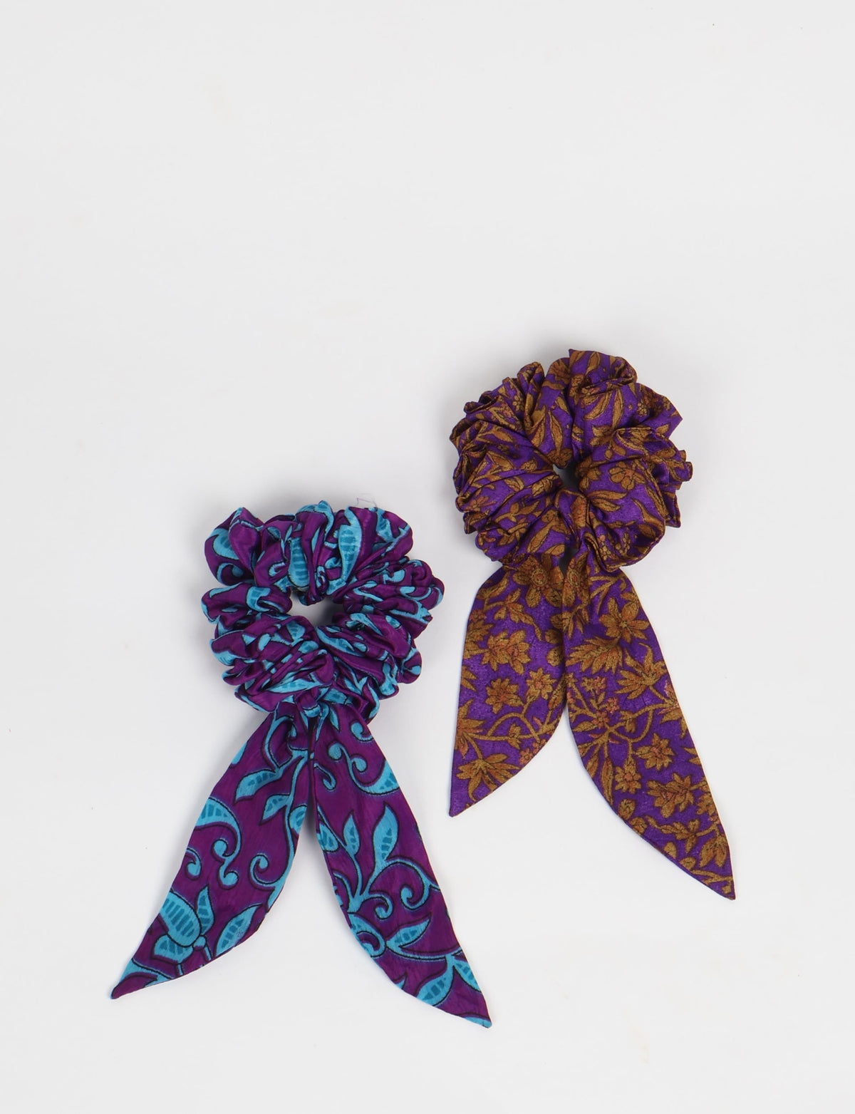 Set of two fabric scrunchies with attached tails, one purple with blue abstract pattern, the other purple with brown floral print