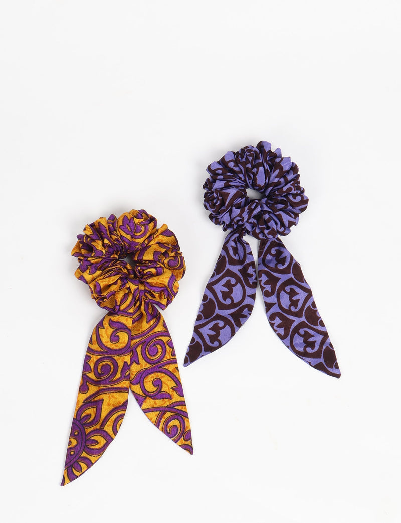 Set of two abstract patterned scrunchies with attached tails, one in purple and brown, the other in yellow and purple, flat lay