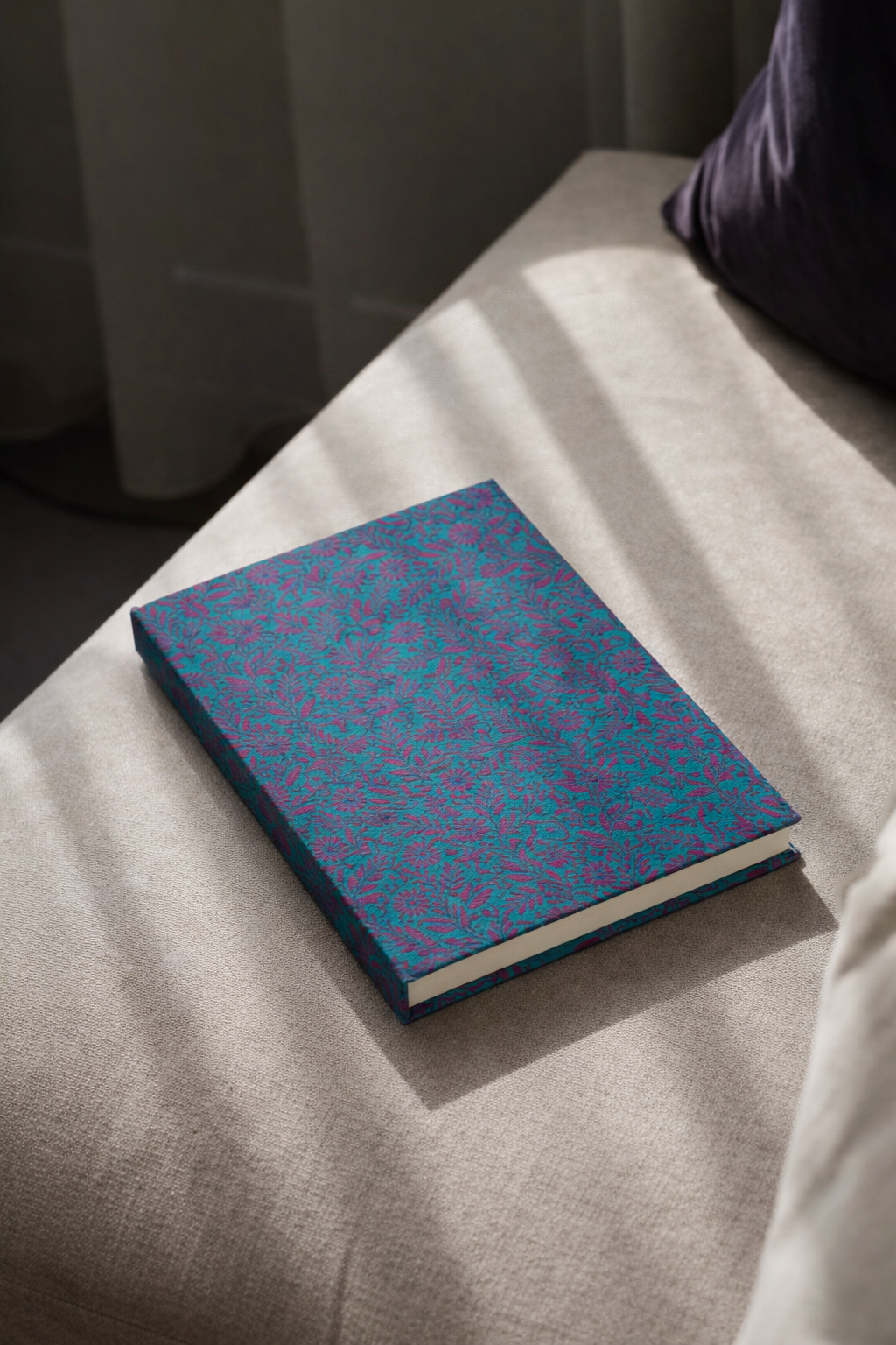 A5 hard cover notebook with blue and purple tropical floral pattern lying on beige fabric surface in natural light