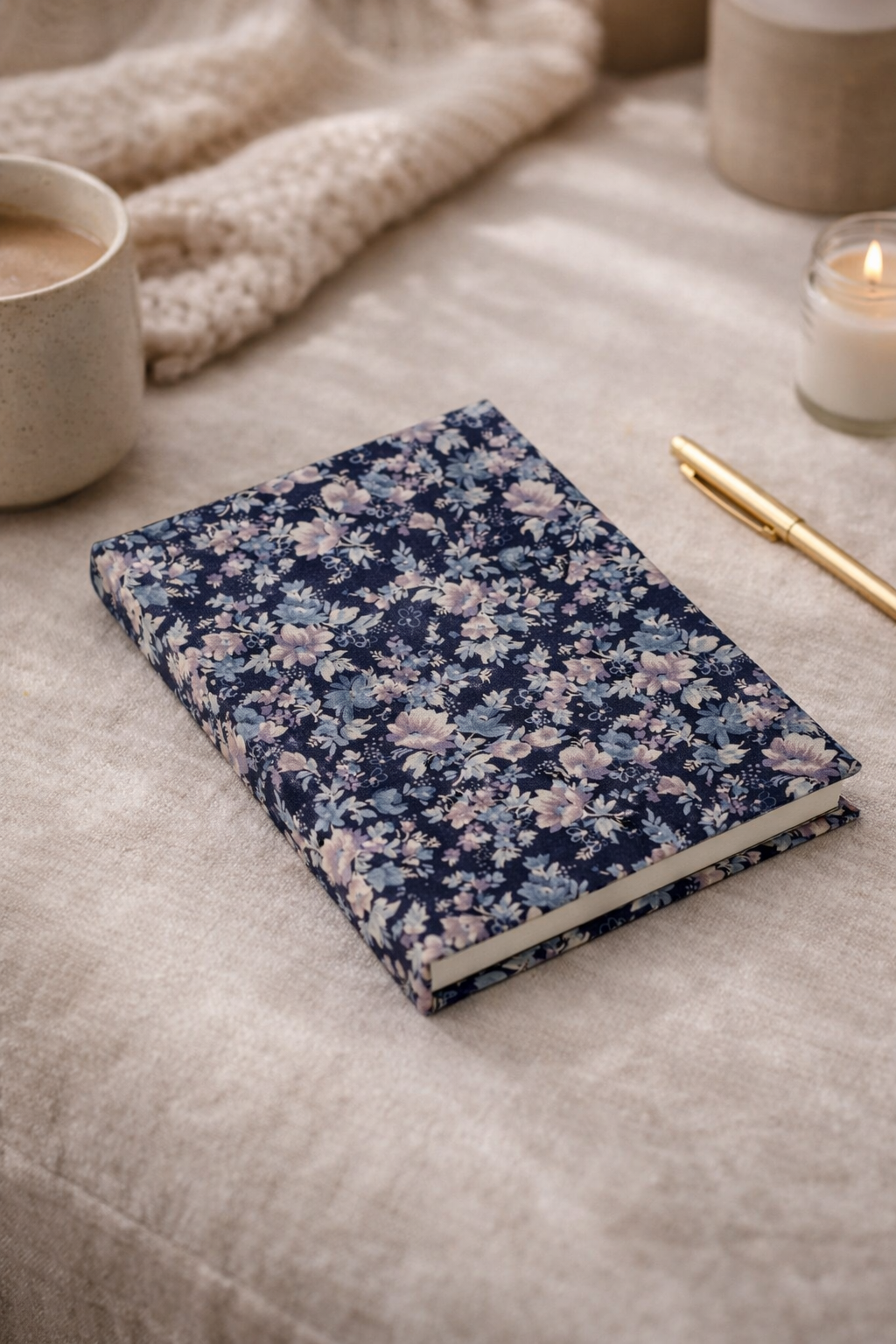 A5 hard cover notebook with blue floral pattern lying on beige fabric next to a gold pen, candle, and coffee cup