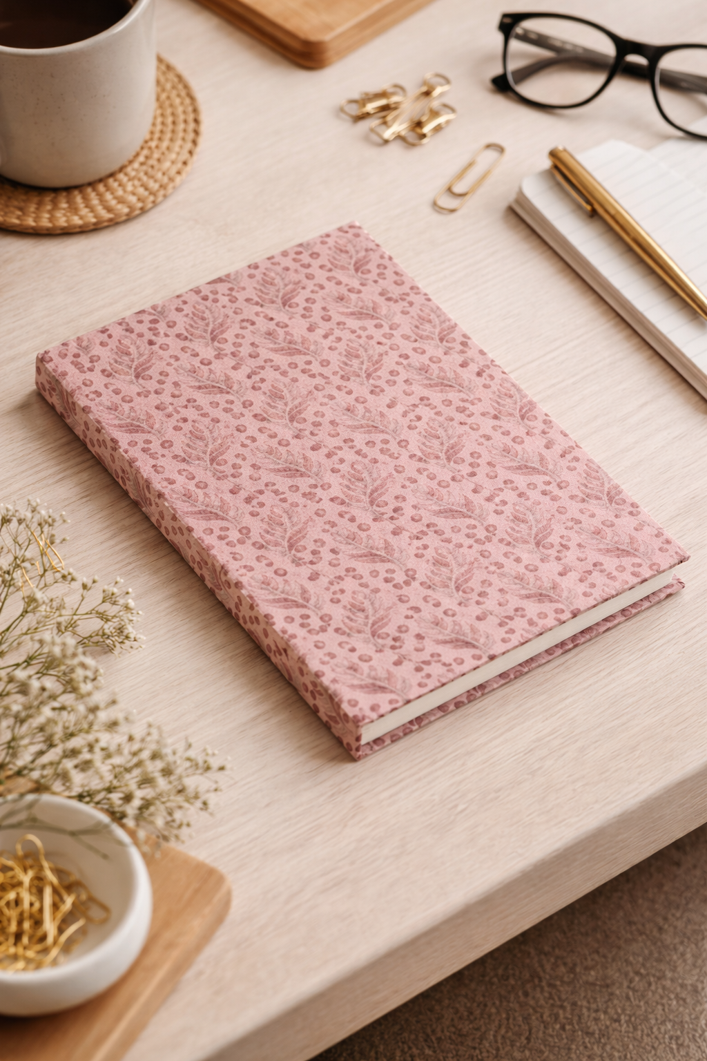 A5 hard cover notebook with pink leaf and dot pattern, shown closed on a light wood desk with office accessories