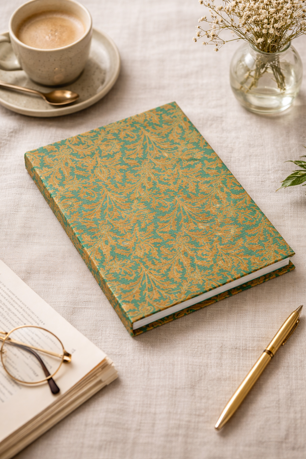 A5 hard cover notebook with green and gold leaf pattern, shown flat on beige fabric with coffee, glasses, and pen nearby