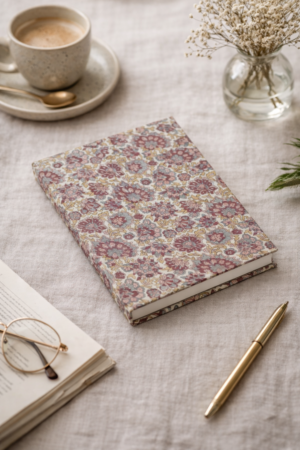 A5 hard cover notebook with beige floral pattern lying on a linen cloth beside a gold pen and eyeglasses on an open book