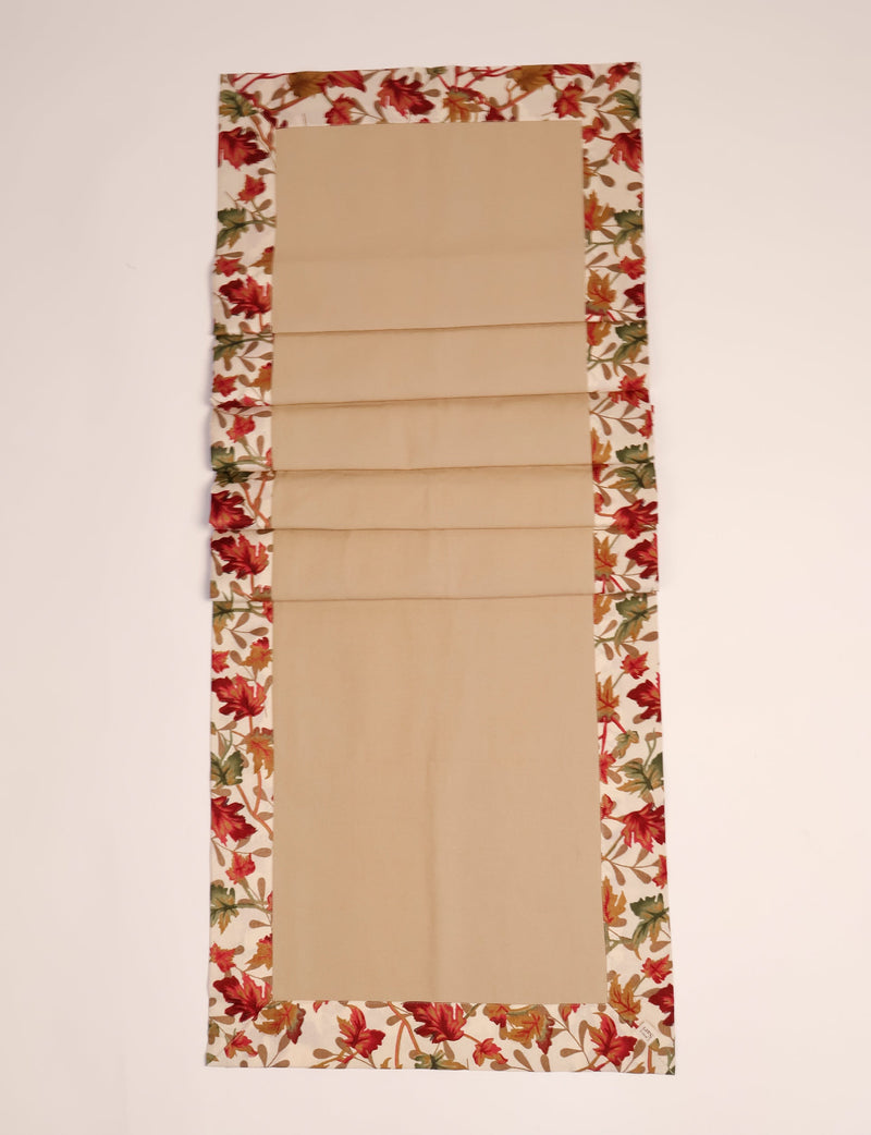 Beige fabric table runner with a border of red, orange, and green autumn leaves on a cream background, flat laid
