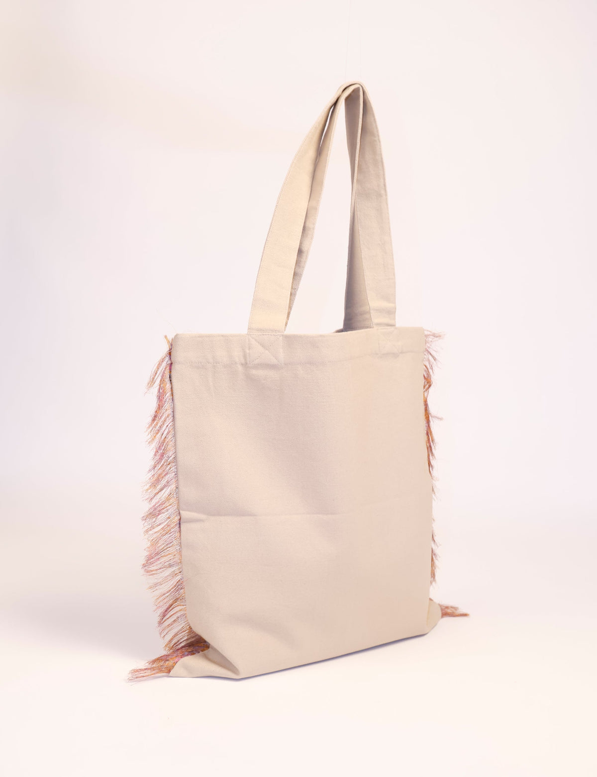 Beige canvas shopper bag with long handles and multicolored fringe detail on the sides, shown hanging upright