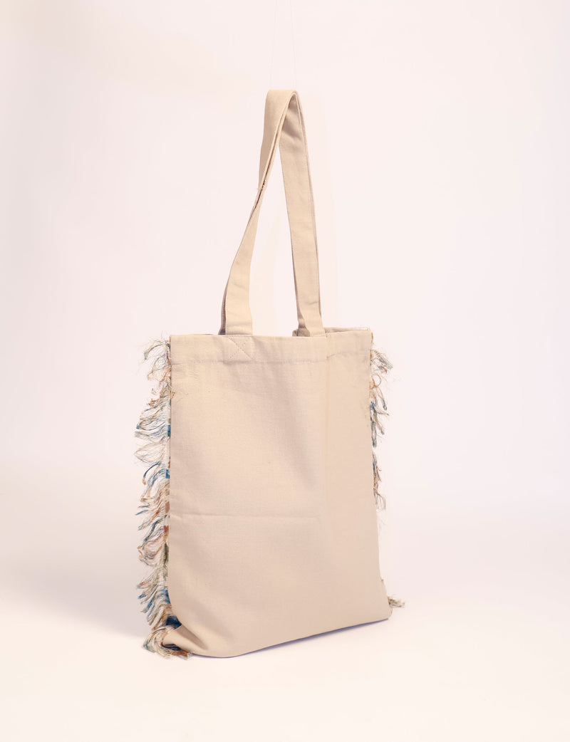 Beige canvas shopper bag with long handles and multicolored fringe detailing on the sides, front view hanging