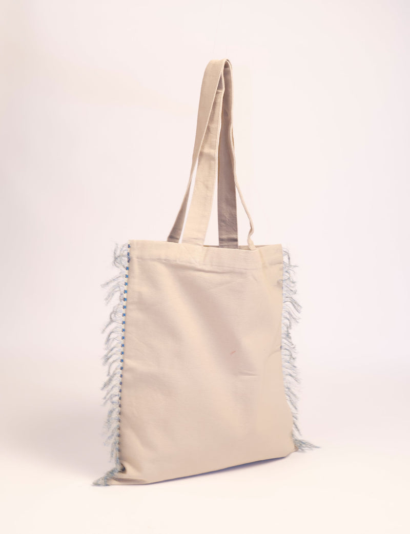 Beige canvas shopper bag with long handles and blue-stitched fringe detail on sides, shown hanging against white background