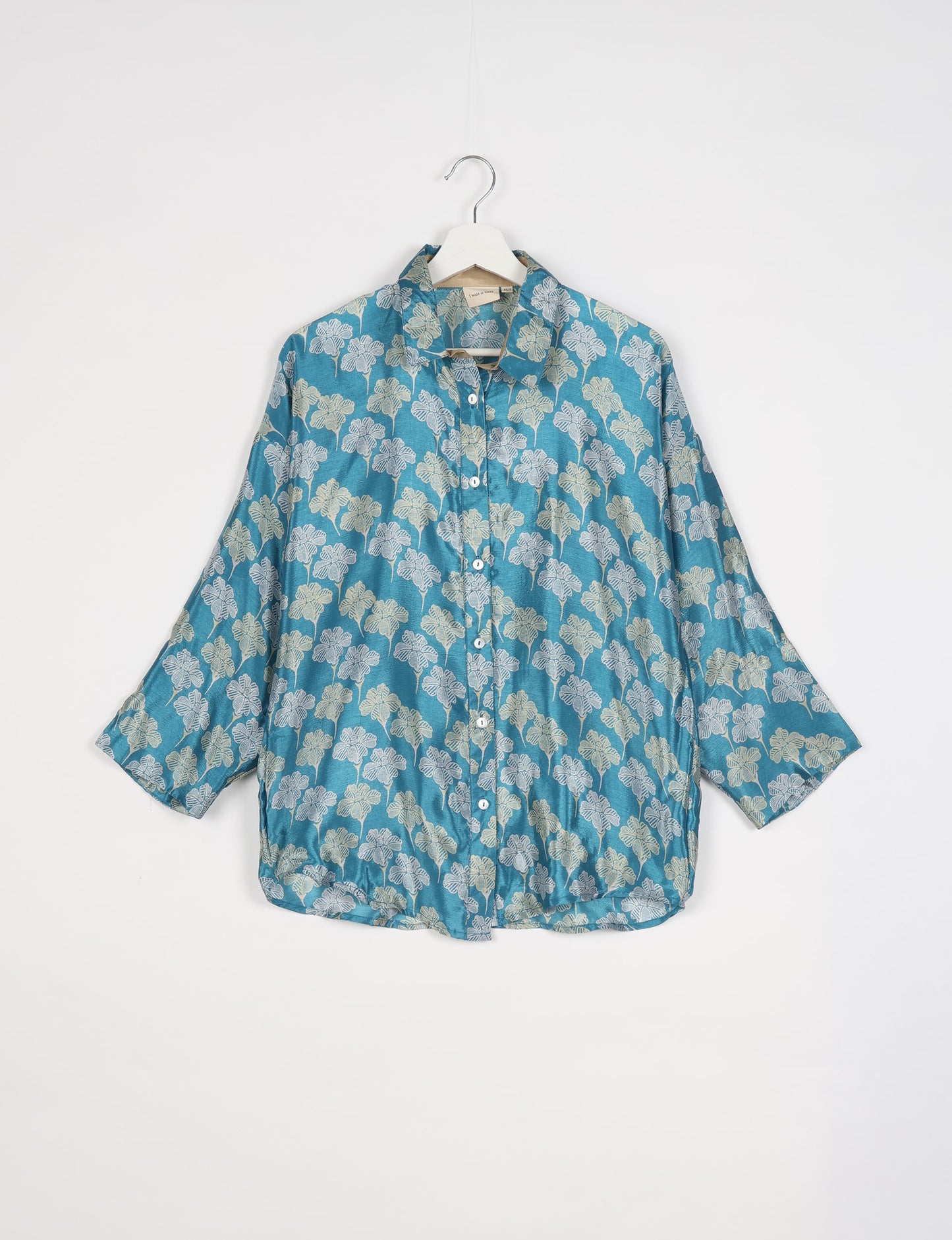Blue oversized button-up shirt with white and beige floral print, spread collar, and three-quarter sleeves, hanging on white
