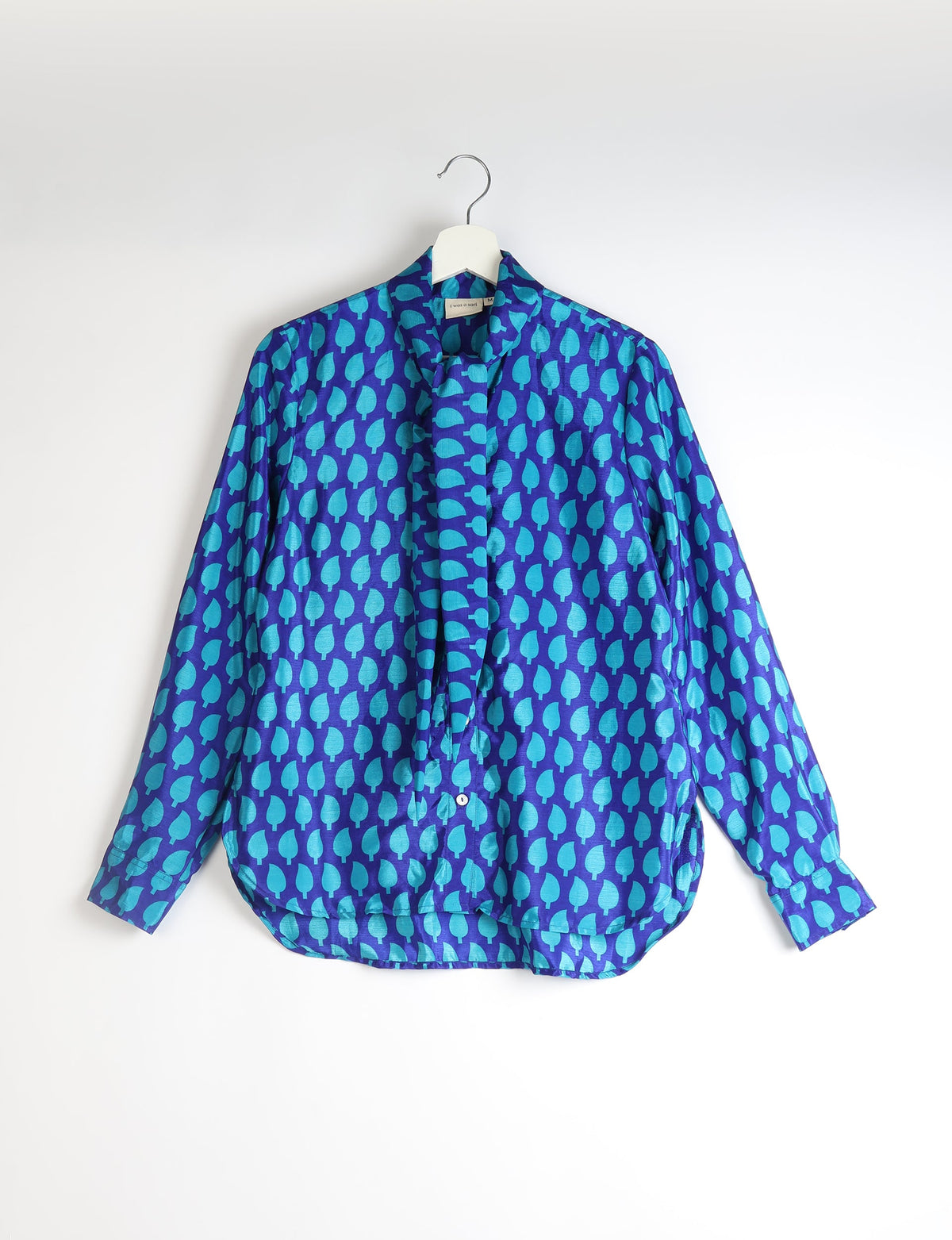Blue long-sleeve button-up shirt with turquoise leaf pattern and attached bowtie, hanging on white hanger against white