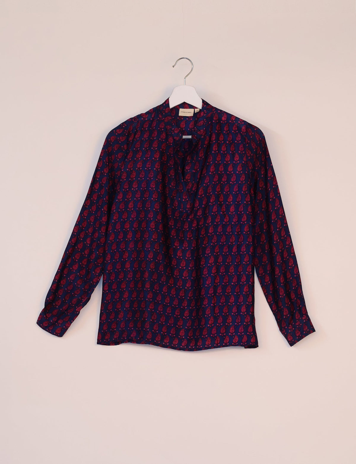 Long-sleeve blue blouse with red paisley micro design, collarless neckline, hanging on white hanger against pale background
