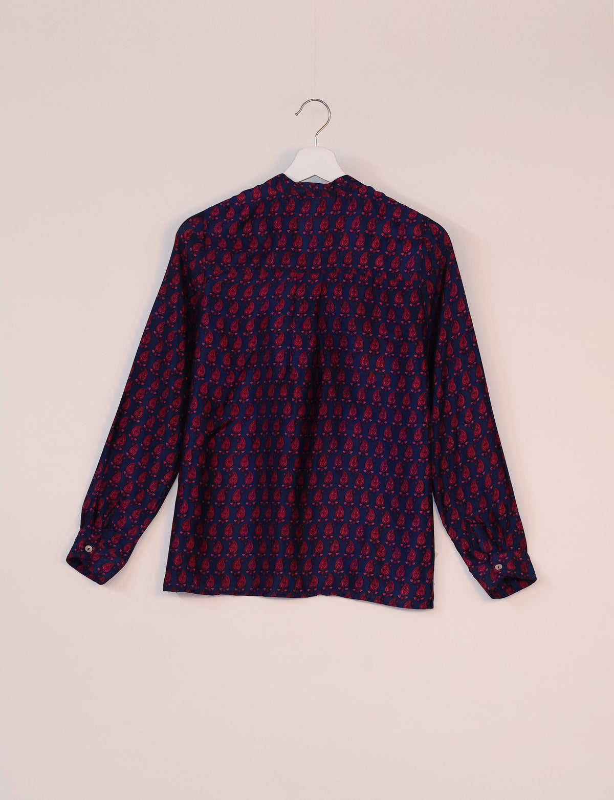 Blue blouse with red paisley micro design, long sleeves, buttoned cuffs, shown from back on white hanger against beige wall