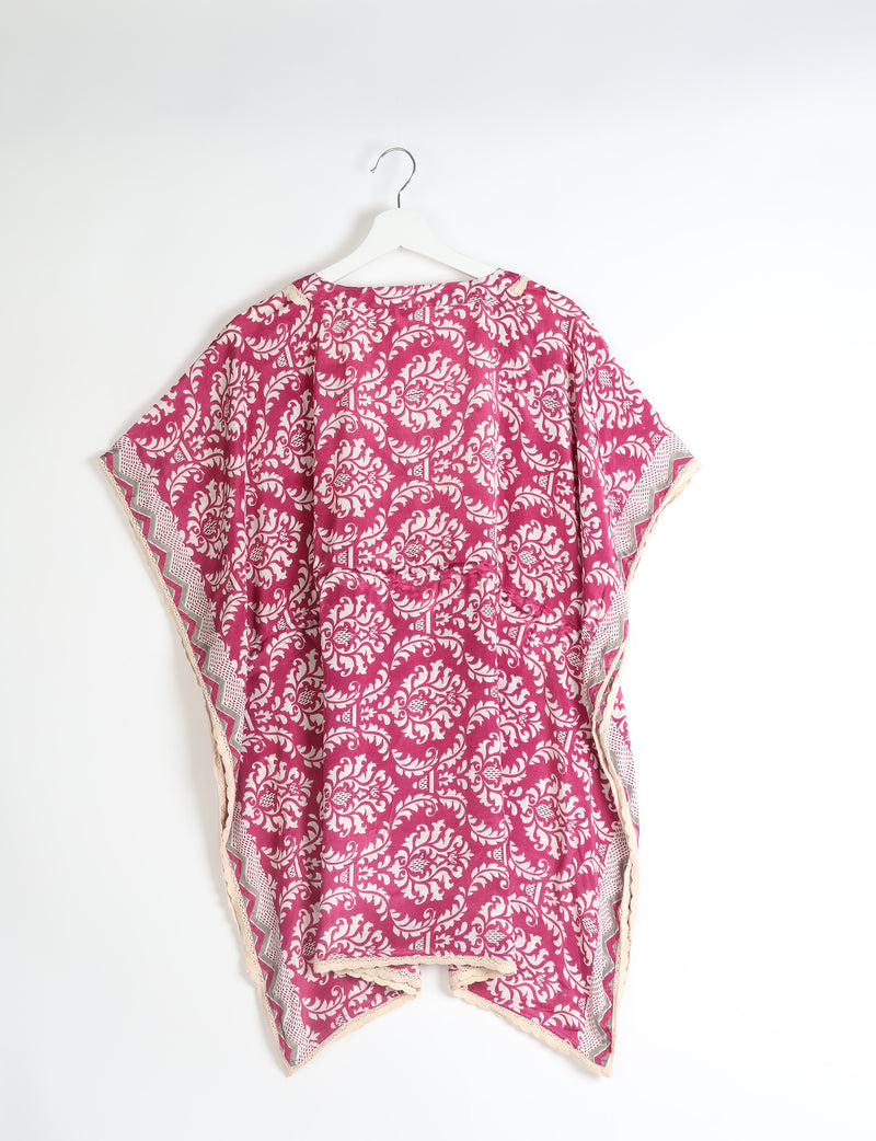 Short pink kaftan with white tropical floral pattern and lace trim hanging on white hanger against white background
