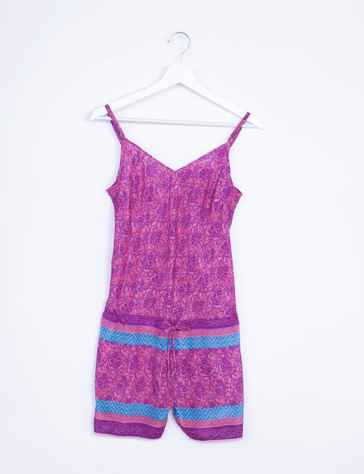 Pink tropical playsuit with lace trim, adjustable straps, and blue and purple patterned bands, hanging on a white hanger