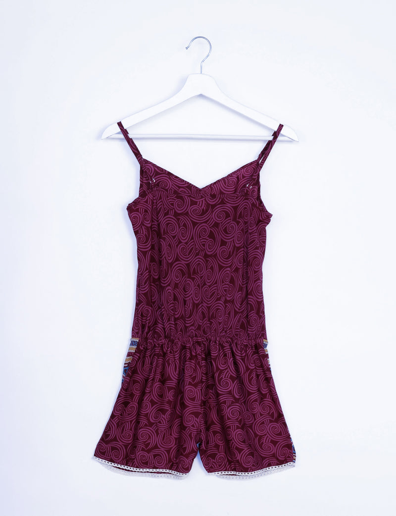 Red playsuit with abstract swirl pattern, adjustable spaghetti straps, elastic waist, and white lace trim on shorts, hanging flat