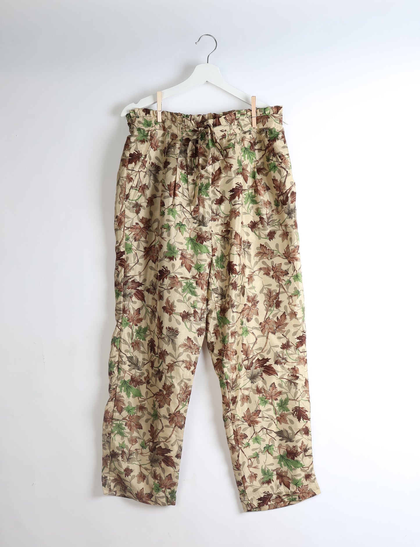Beige paperbag high-waisted pull-on pants with green and brown leaf print, front pockets, and waist tie, hanging on white hanger