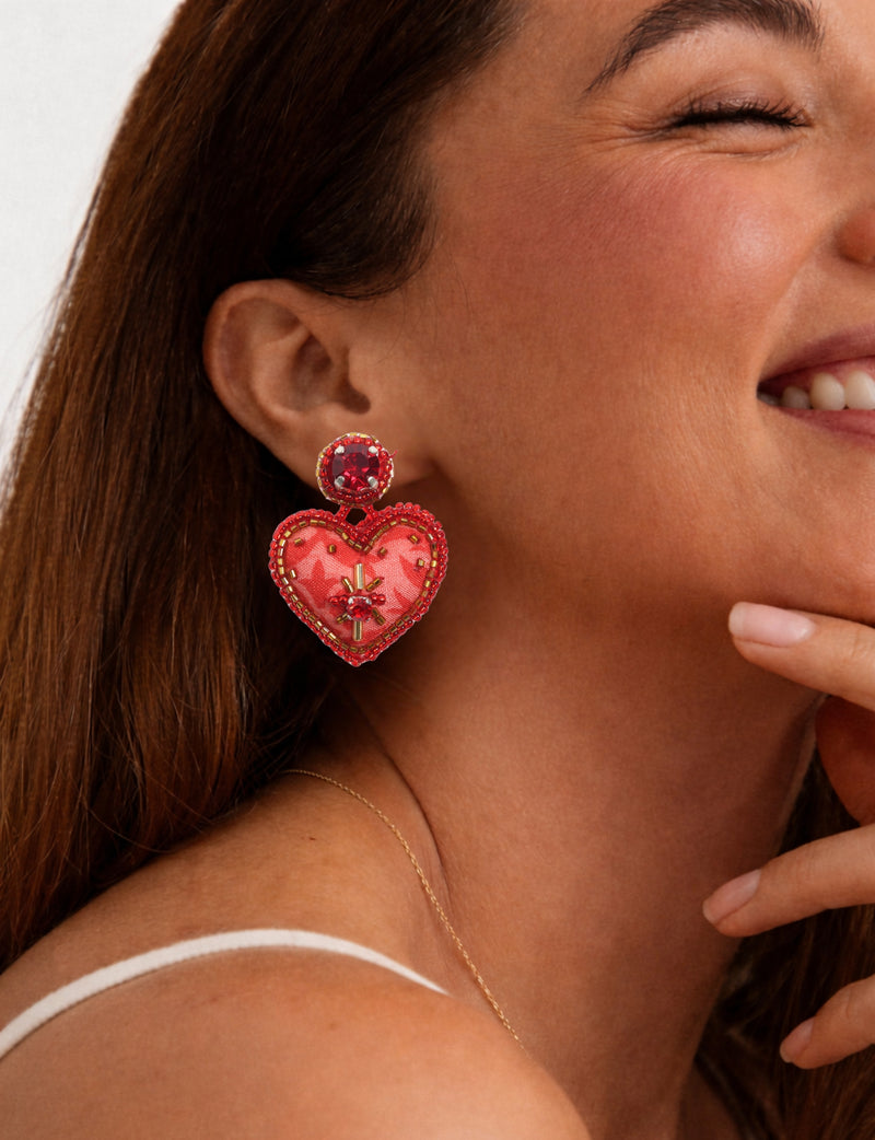 Red heart drop earring with bead and gem embellishments worn by smiling woman in white strap top, side view