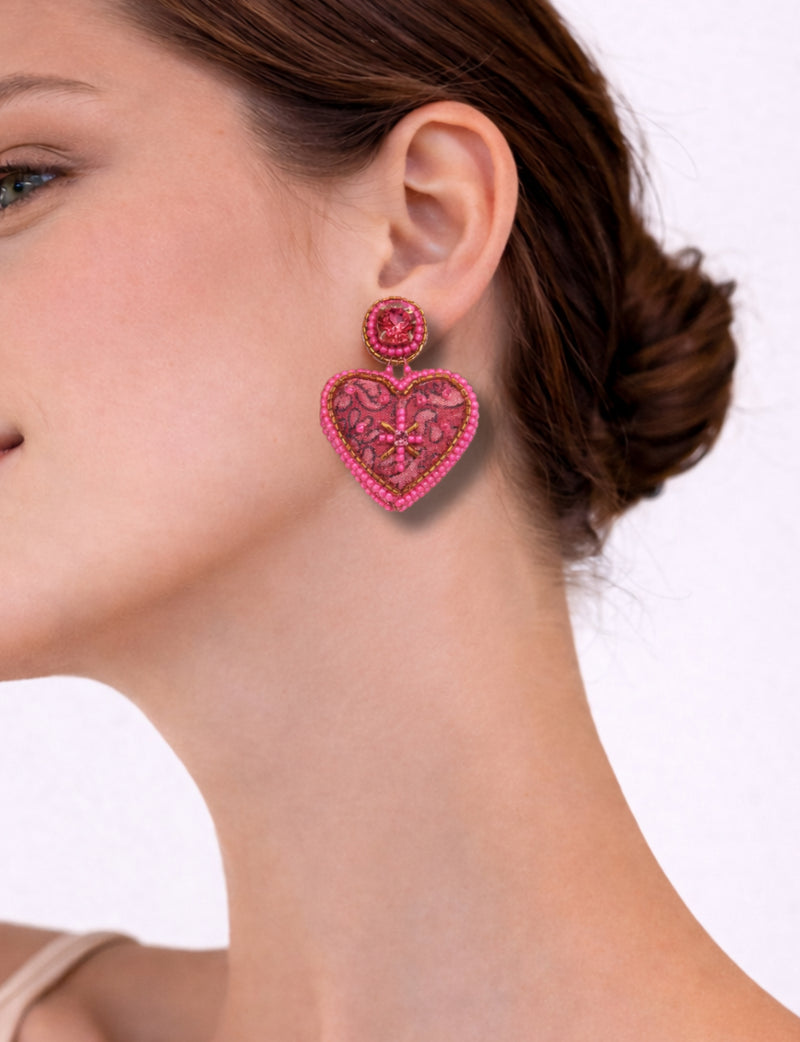 Pink tropical heart drop earrings with bead and lace details worn by a woman with brown hair in a bun, side view