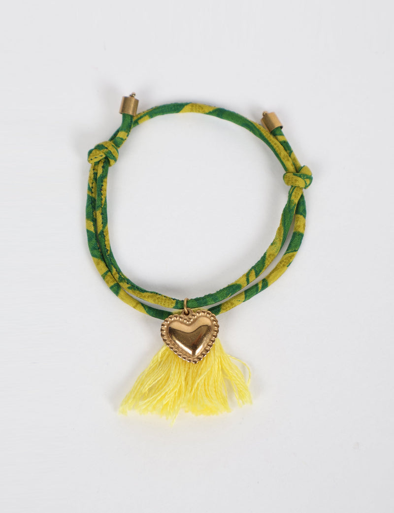 Green and yellow abstract patterned rope bracelet with gold heart charm and yellow tassels, flat lay on white background