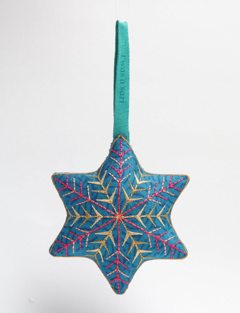Blue fabric snowflake ornament with gold and pink embroidered details hanging by a teal loop against white background