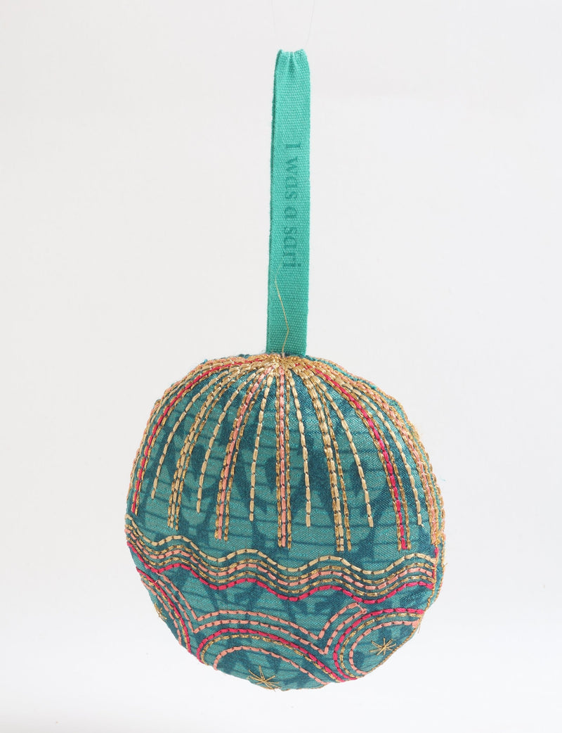 Round fabric ornament in green with gold and pink stitched patterns, hanging by a green fabric loop with text