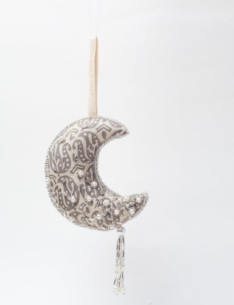 Silver crescent moon ornament with paisley pattern, pearl and bead embellishments, hanging by beige fabric loop with beaded tassel