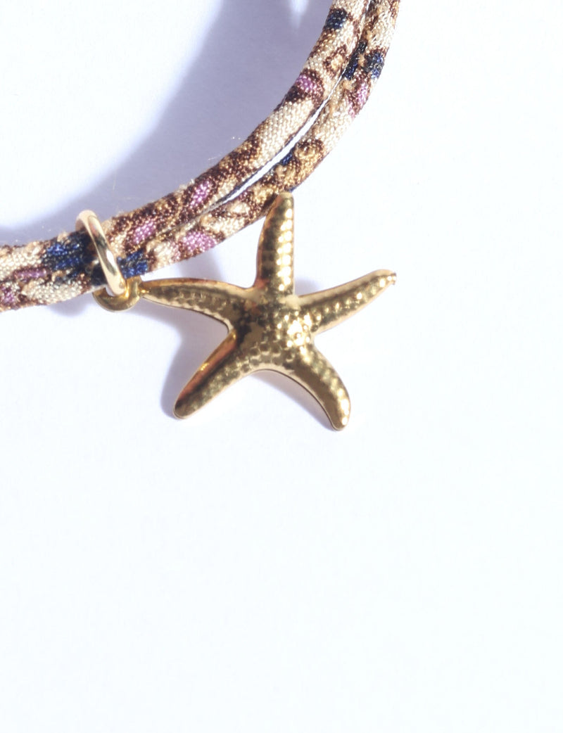 Close-up of a cord bracelet with a gold starfish charm and a patterned fabric cord in beige, brown, blue, and purple tones
