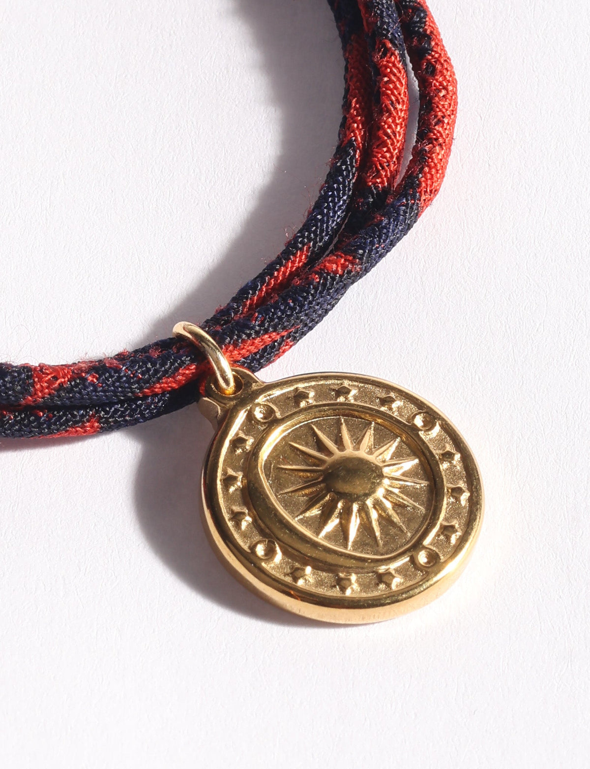 Blue and red braided cord bracelet with a round gold sun medallion charm shown in close-up on white background