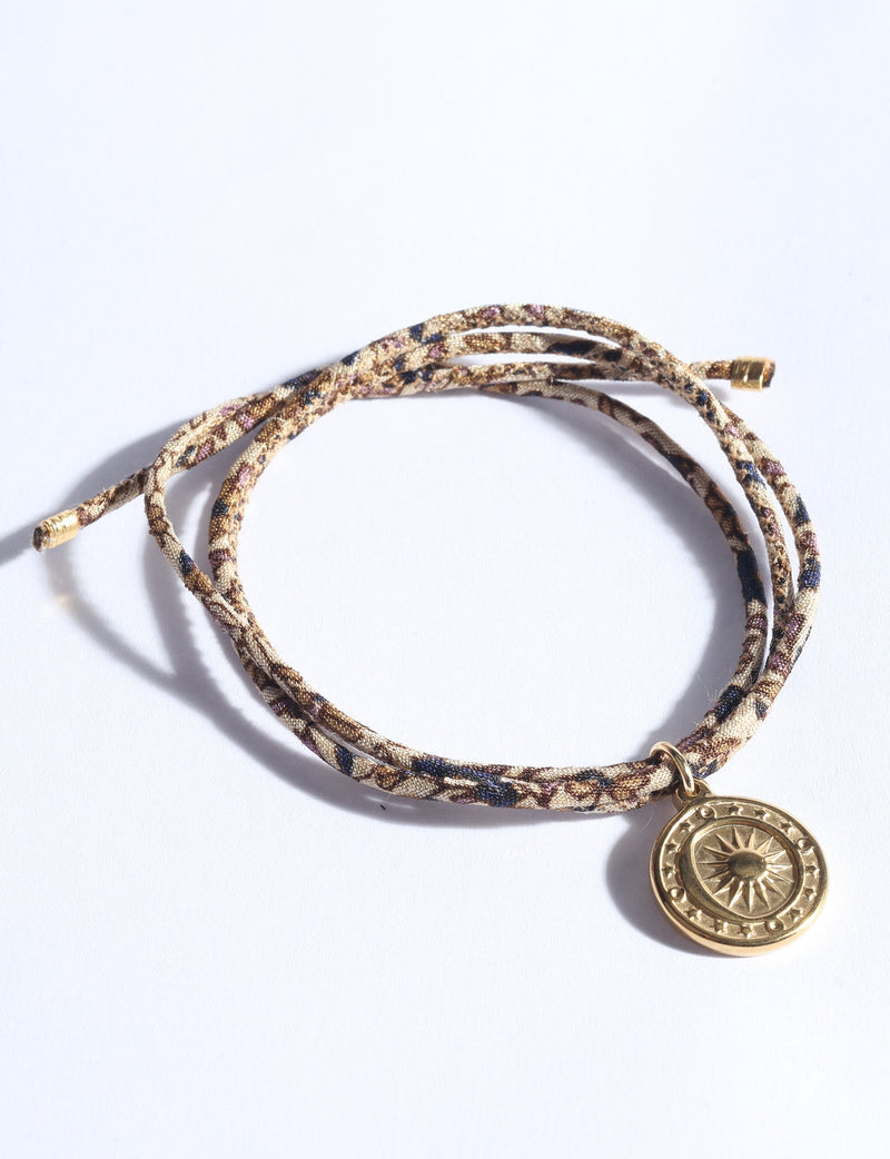 Beige abstract patterned cord bracelet with gold-tone sun medallion charm, shown flat on white background