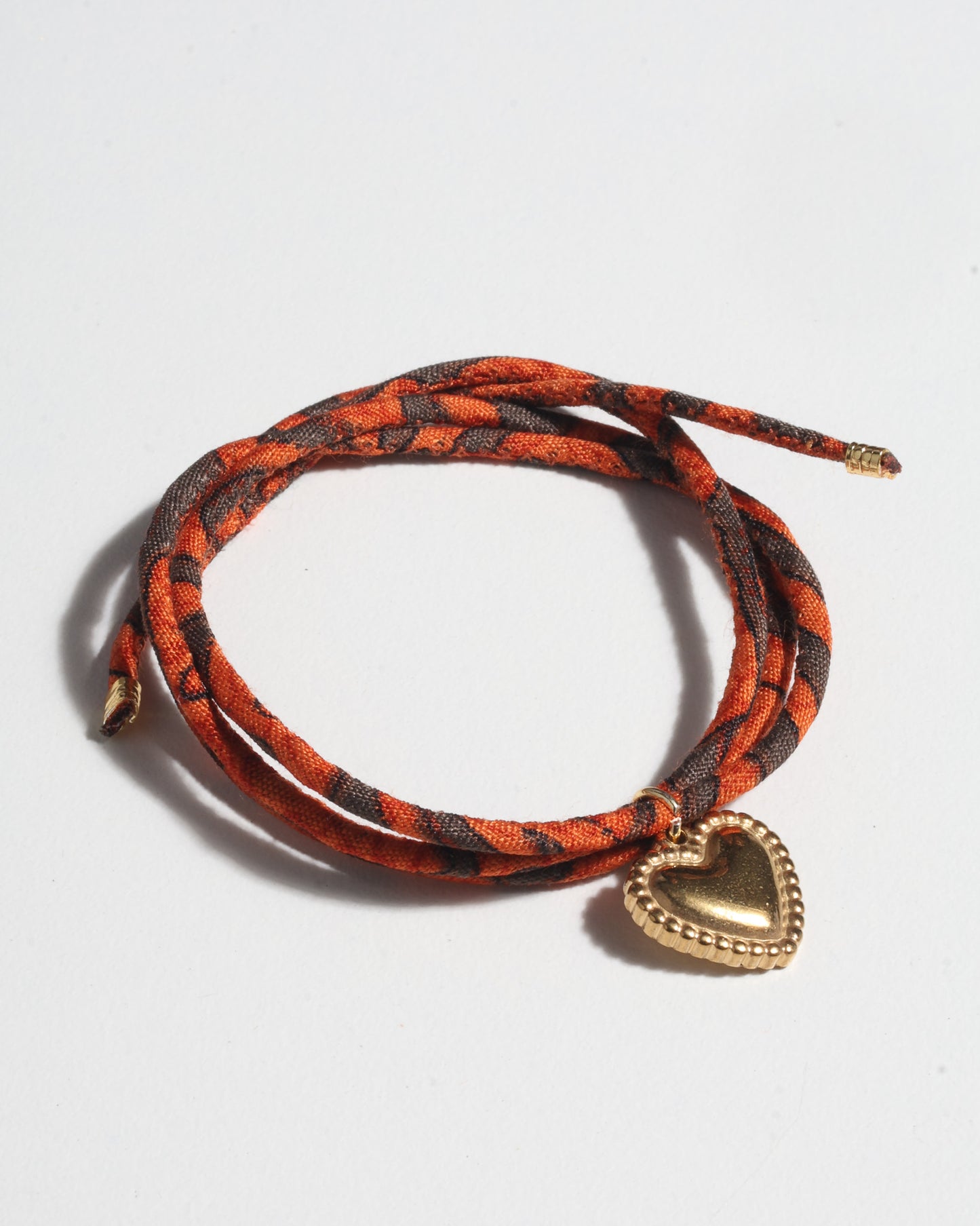 Orange and dark brown patterned cord bracelet with gold-tone heart charm, shown coiled on white background