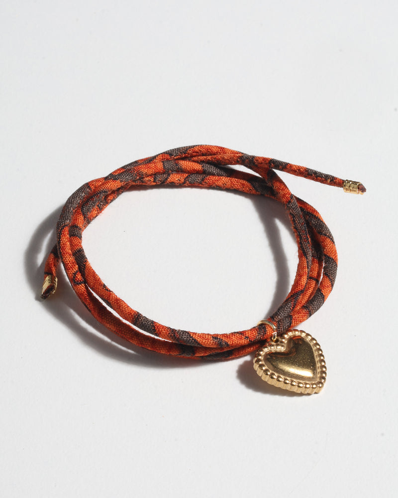 Orange and dark brown patterned cord bracelet with gold-tone heart charm, shown coiled on white background