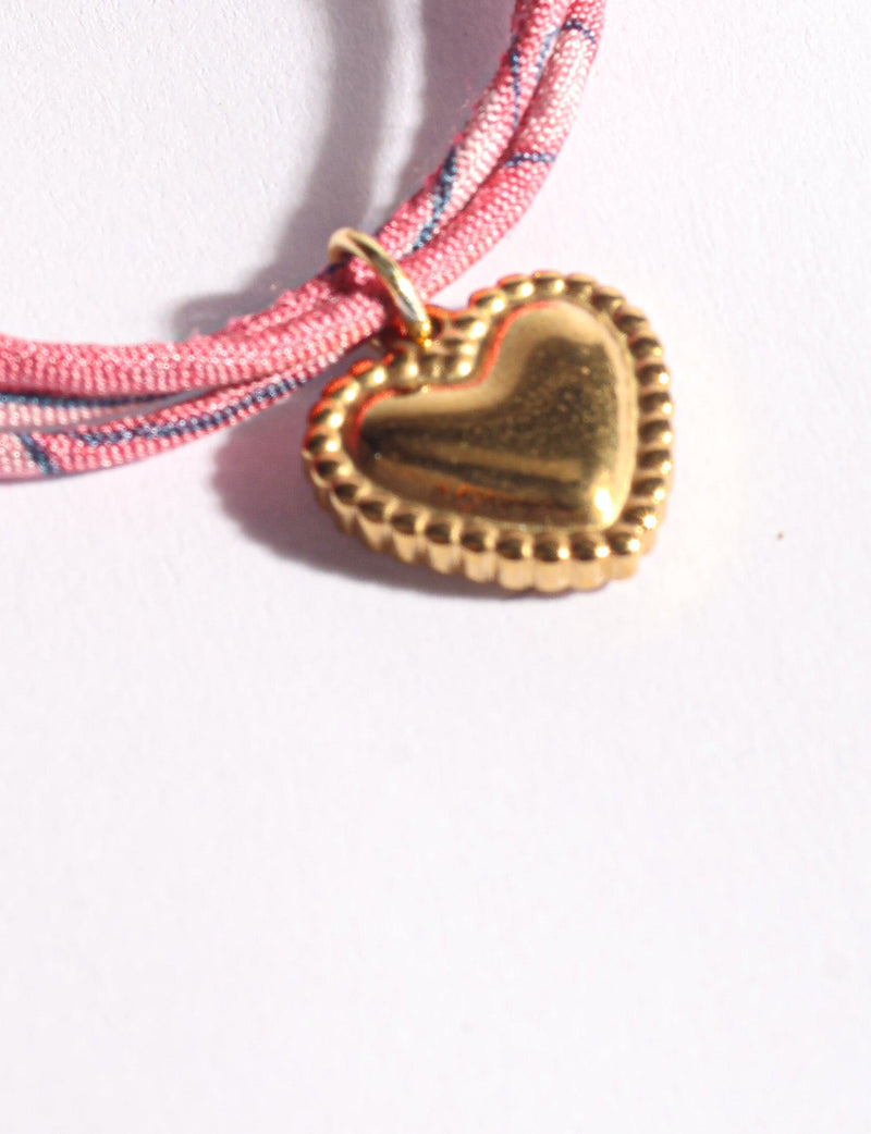 Pink cord bracelet with blue accents and a gold-tone heart-shaped charm with scalloped edges, close-up view