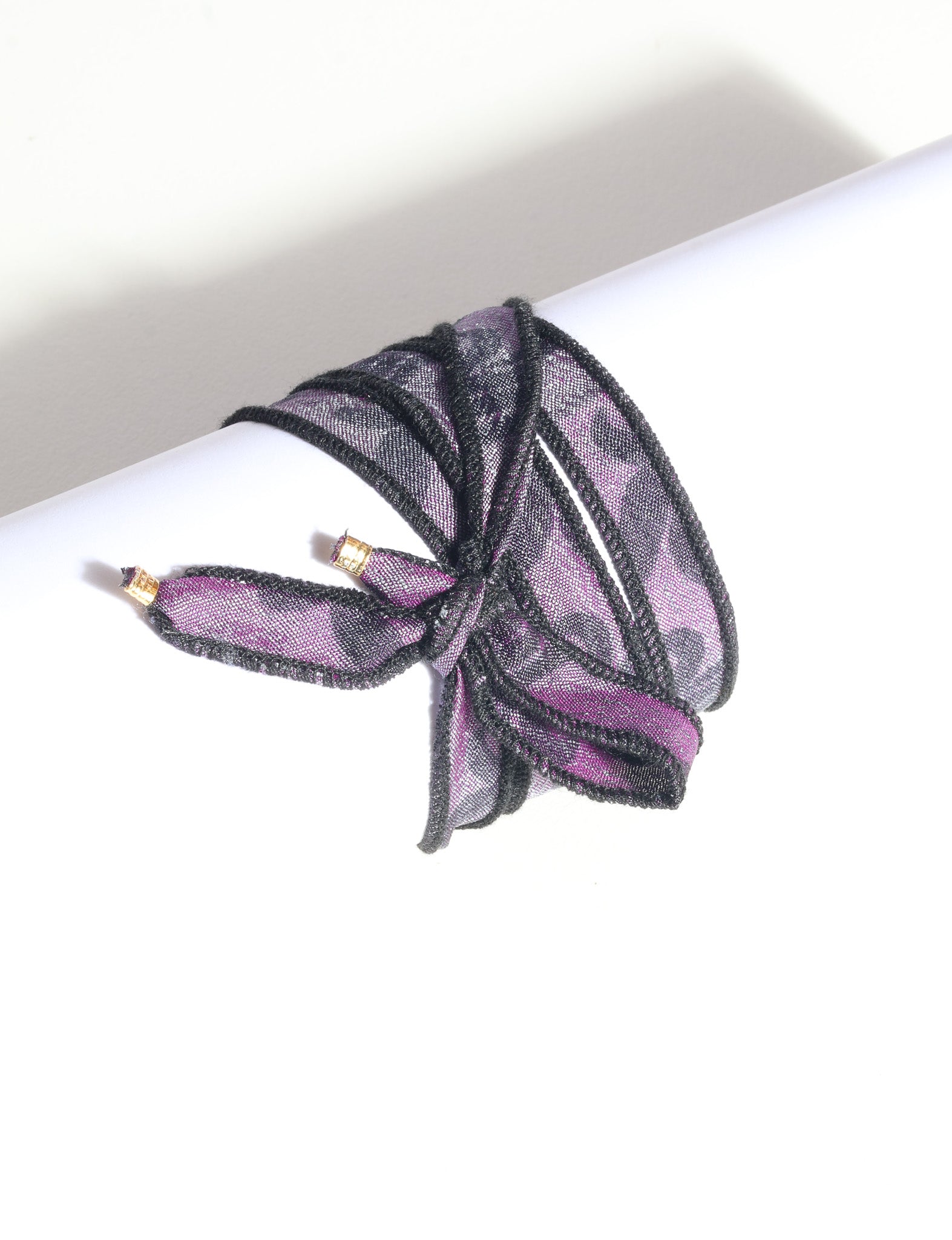 Purple and black wrap-around fabric bracelet with gold-tone charm ends, shown tied on a white cylindrical display