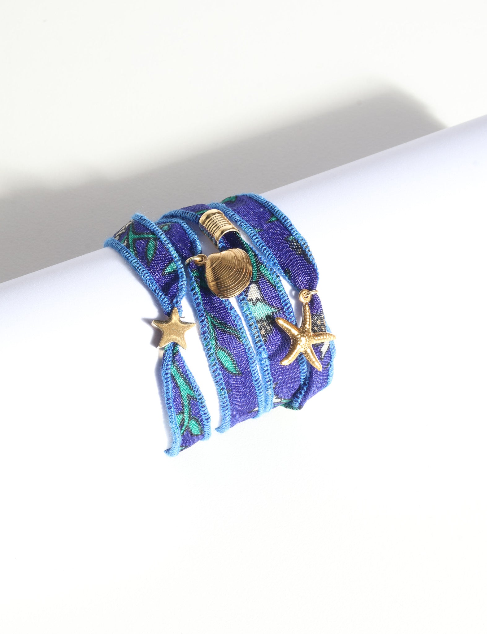 Blue fabric wrap-around bracelet with green and white floral pattern and gold starfish and shell charms, flat lay view