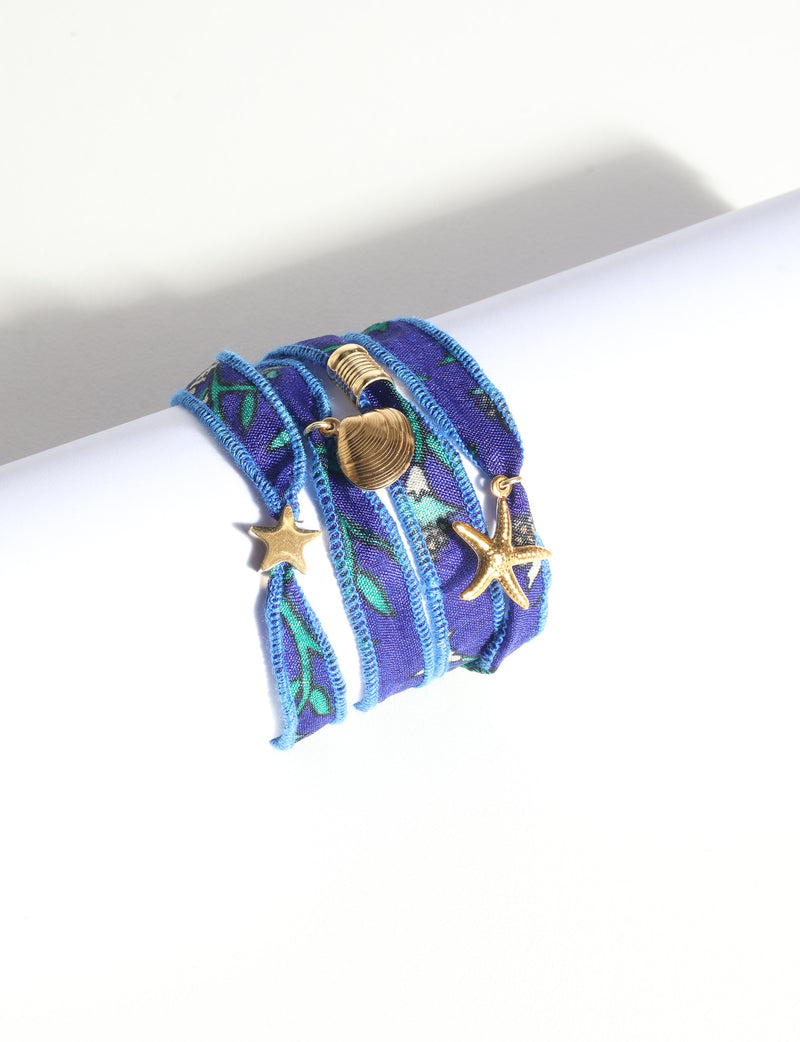 Blue fabric wrap-around bracelet with green and white floral pattern and gold starfish and shell charms, flat lay view