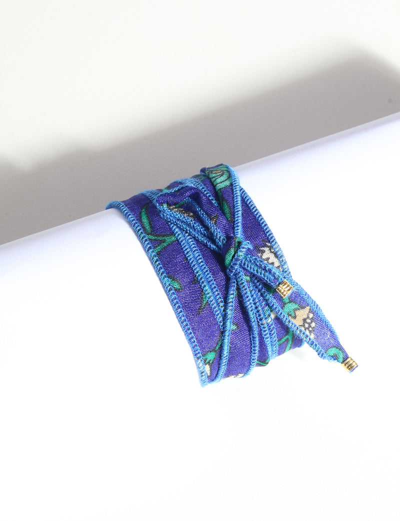 Blue fabric wrap-around bracelet with green and white floral pattern and gold-tone charm ends, shown flat on white surface