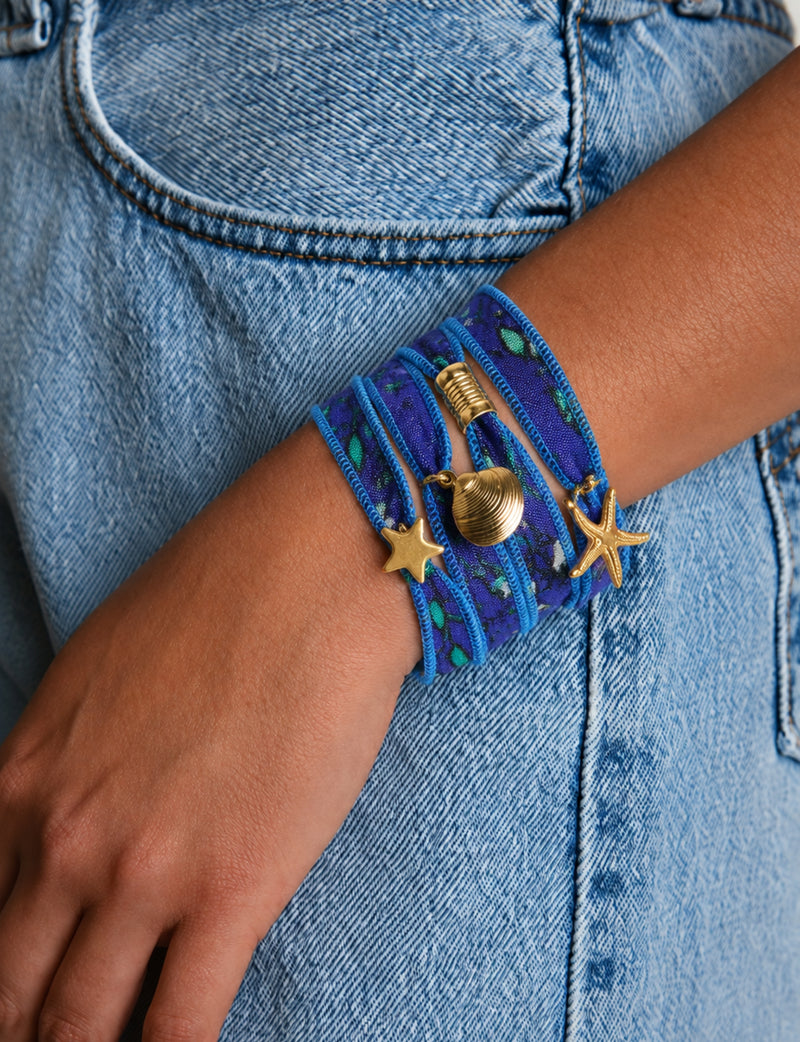 Blue wrap-around fabric bracelet with gold starfish, shell, and star charms worn on wrist over denim jeans pocket