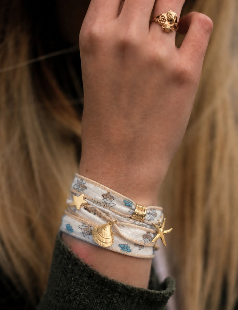 White fabric wrap-around bracelet with blue and brown floral print, gold star, shell, and starfish charms, worn on wrist