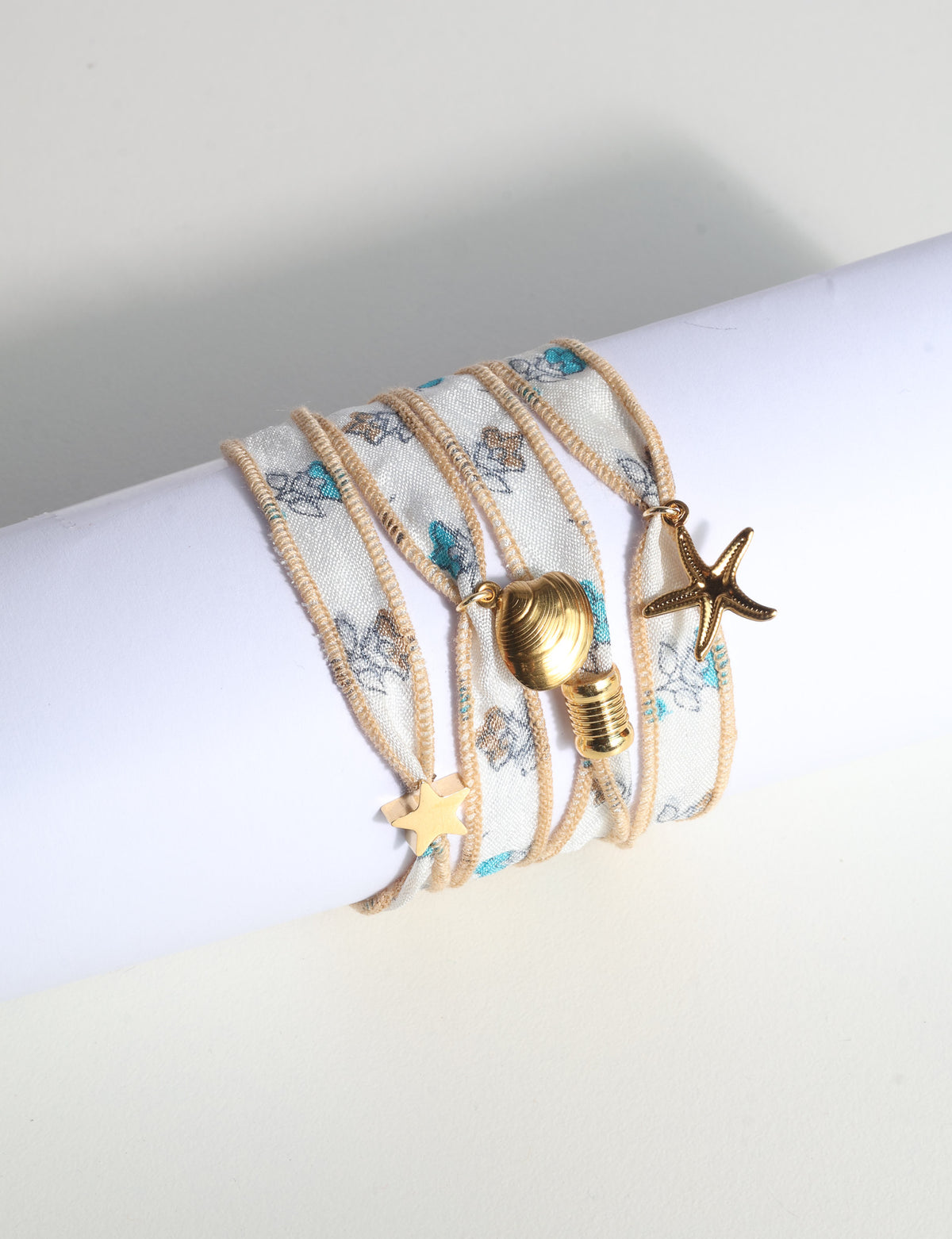 Wrap-around fabric bracelet with beige edges, blue and brown floral print, and gold-tone star, shell, and bead charms