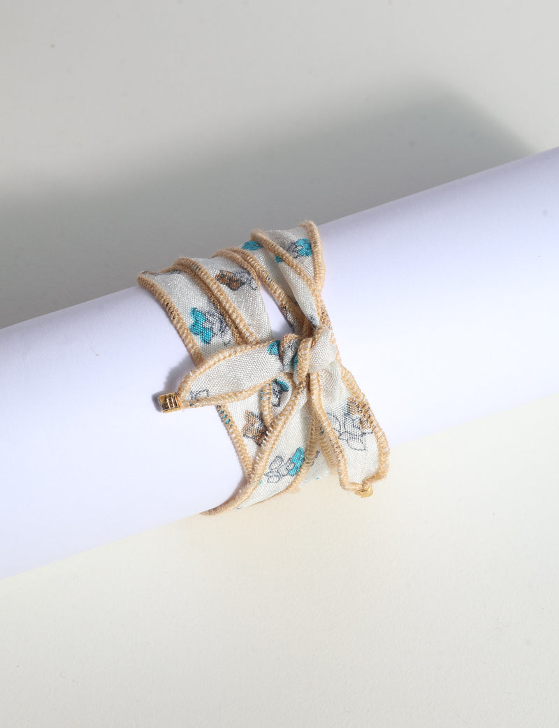 Wrap-around fabric bracelet with beige edges and blue and brown floral print, tied in a bow on white background